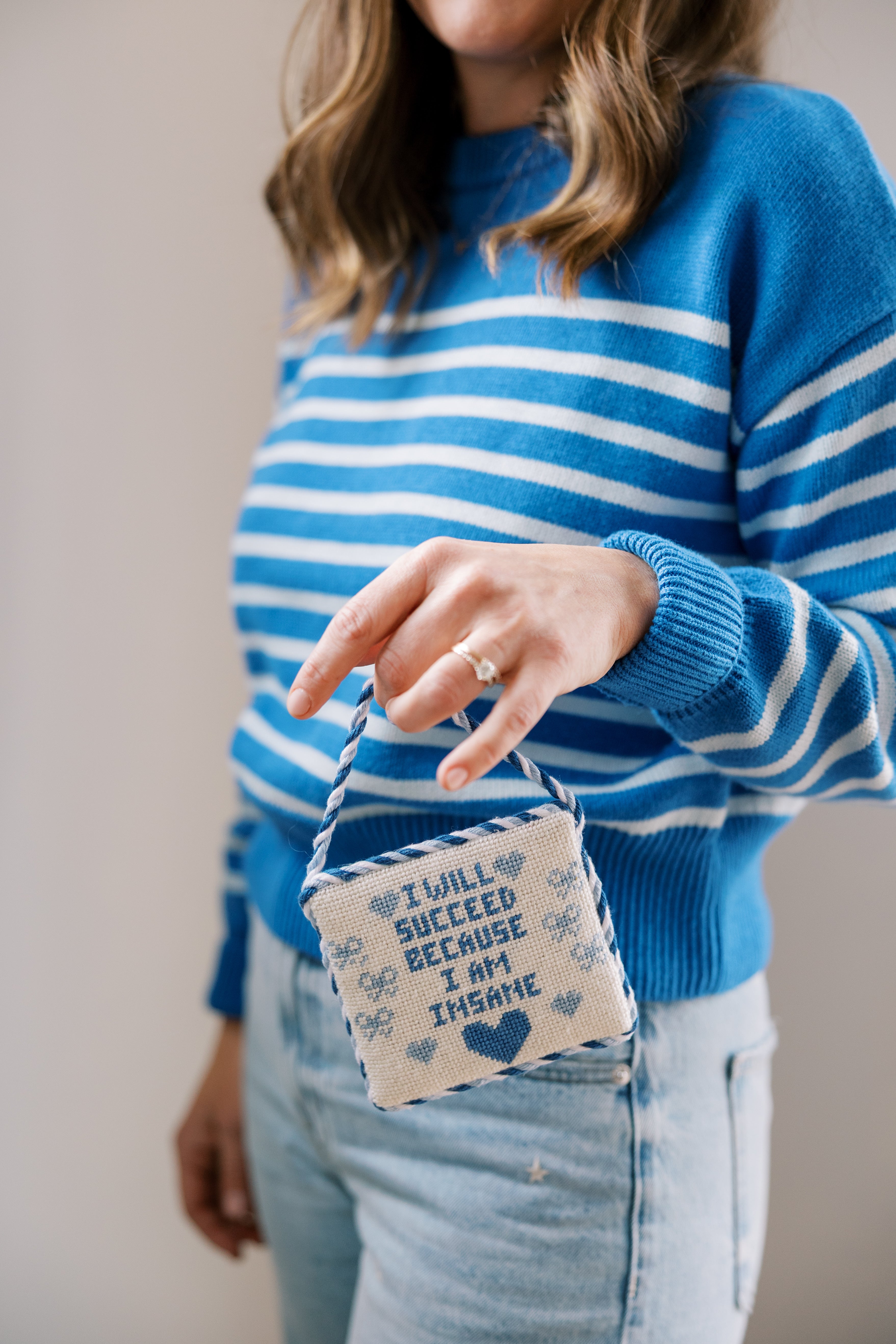 Becca wearing a blue and white striped sweater holding a small finished signs and sayings needlepoint canvas.