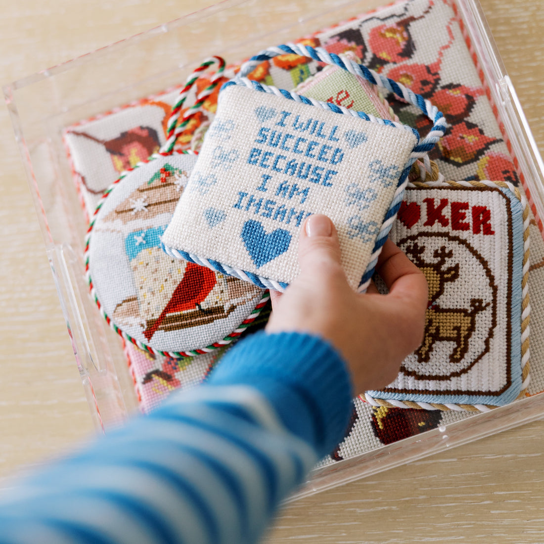 Hand holding a cross-stitched item with text, surrounded by other similar items in a clear plastic case on a wooden surface.