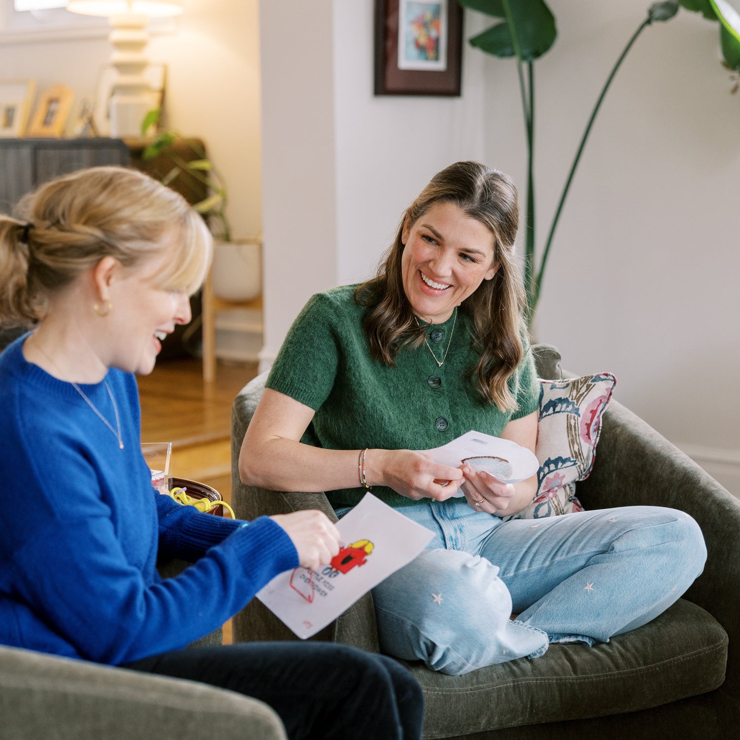 Two women sitting on a couch in a living room, needlepointing together.