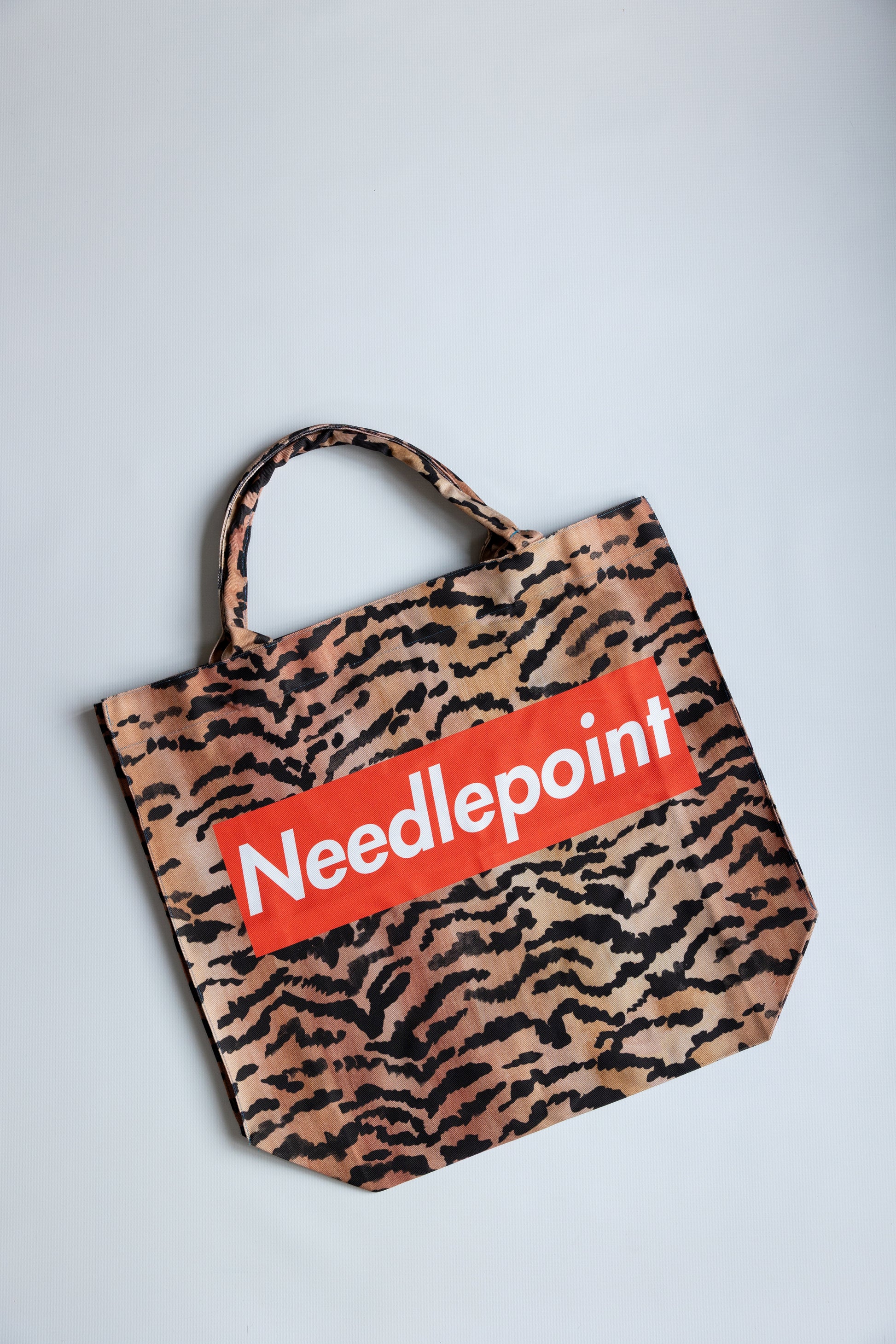 Tote bag with tiger pattern and 'Needlepoint' logo on a red background