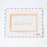 Needlepoint pattern with light pink 'le tired' text on a white background with scallop border