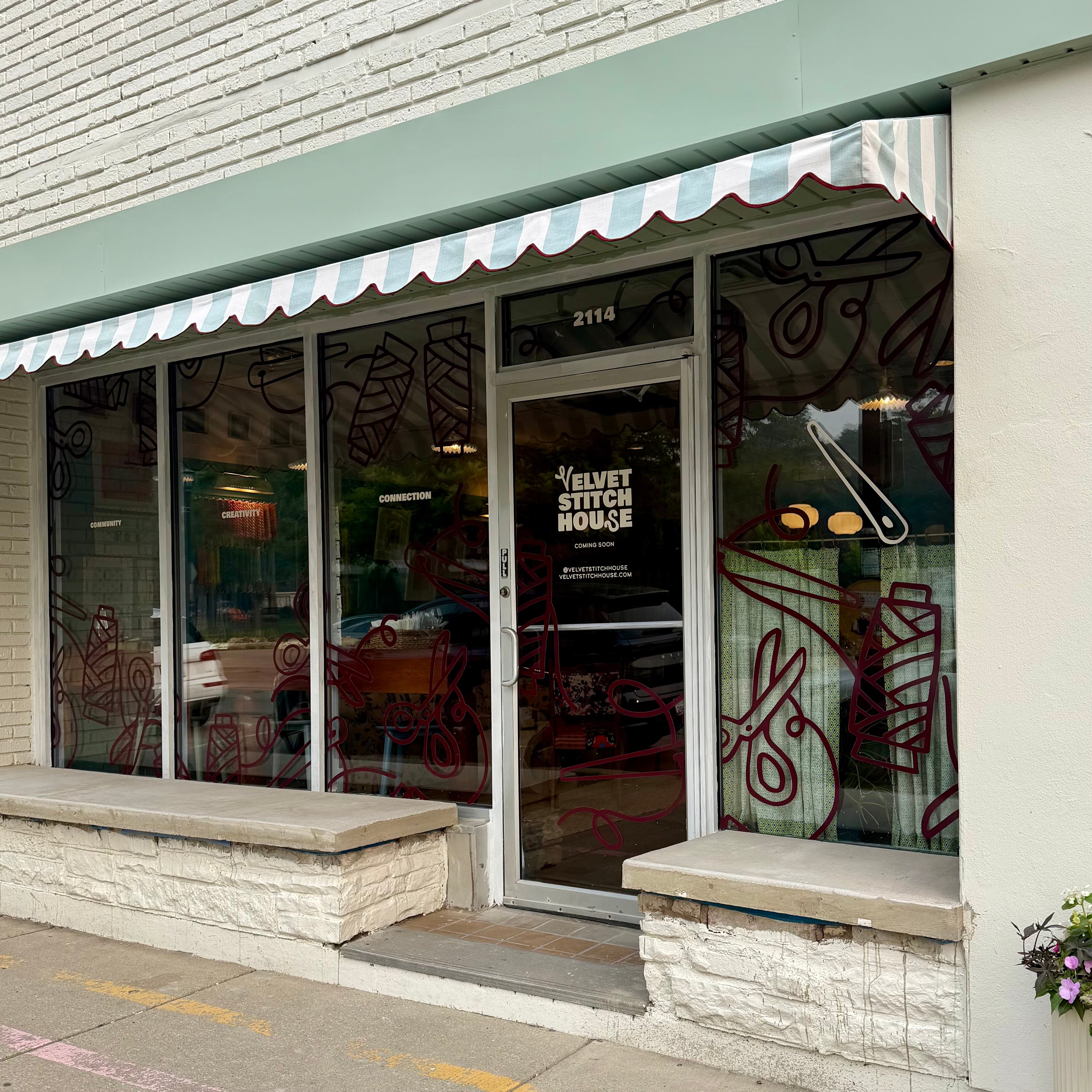 The front entrance of Velvet Stitch House. The shop has a glass door and large windows with a striped awning.