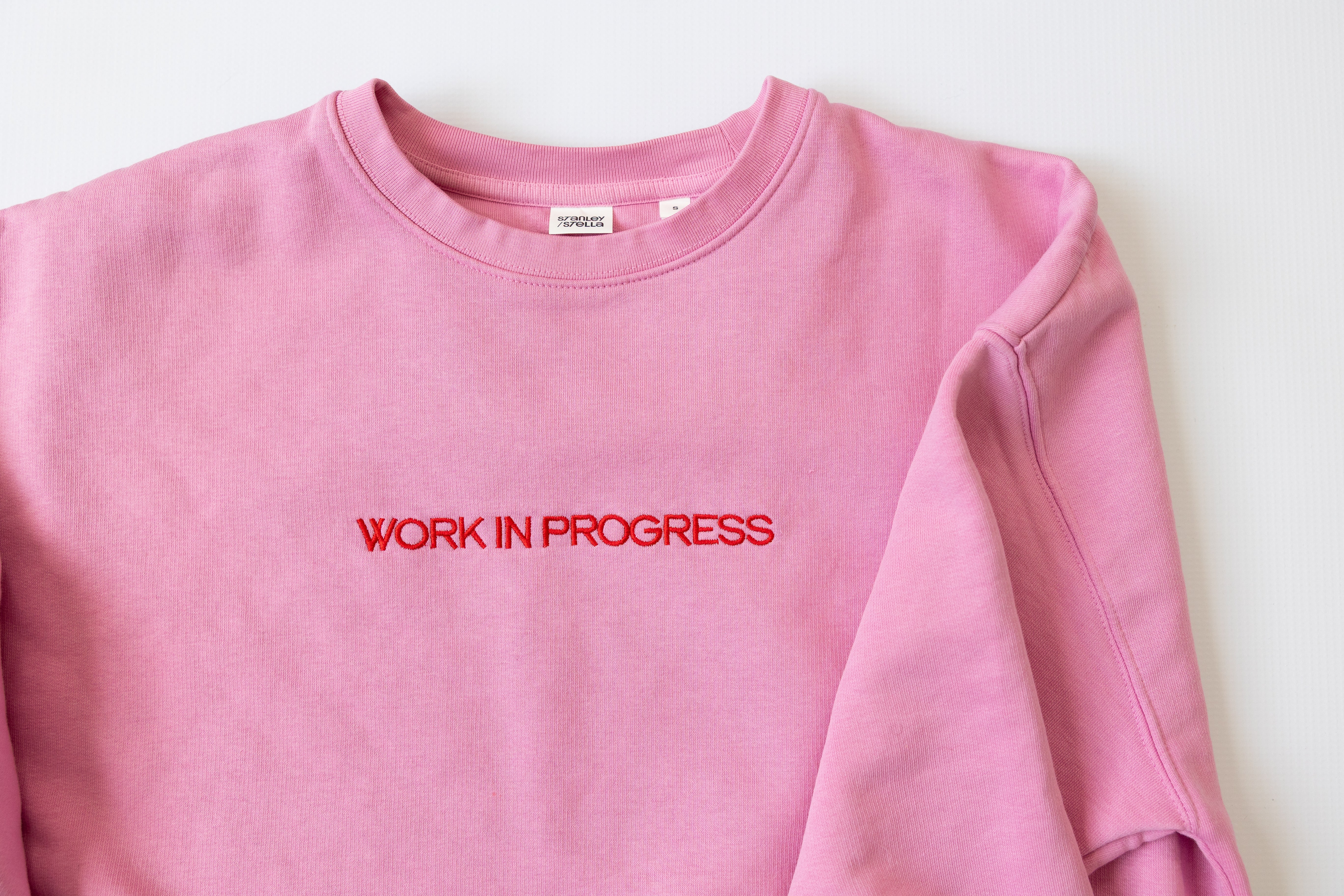 Pink sweatshirt with 'WORK IN PROGRESS' text on a white background