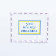 Needlepoint design with 'you are my sunshine' in blue text with a yellow scallop frame on a white background